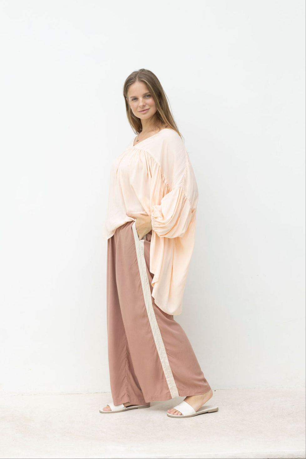 Woman wearing a blush top and wide-leg mocca coloured pants against a white coloured.