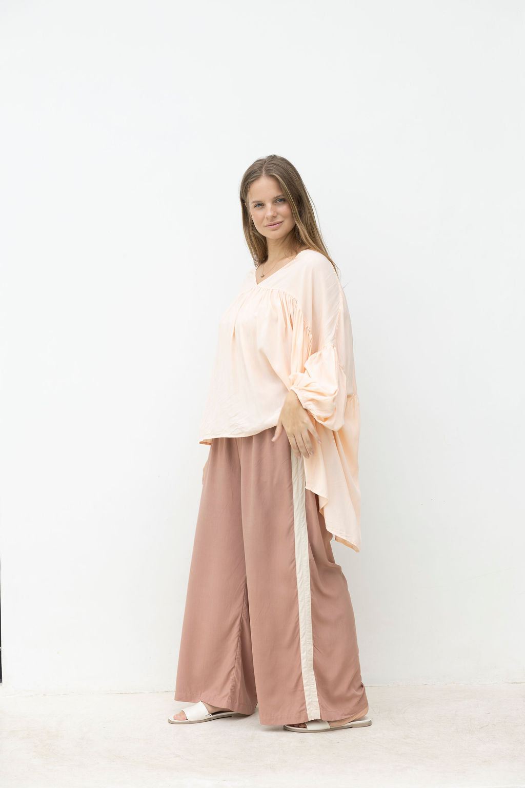 Woman wearing a blush top and wide-leg mocca coloured pants against a white coloured.