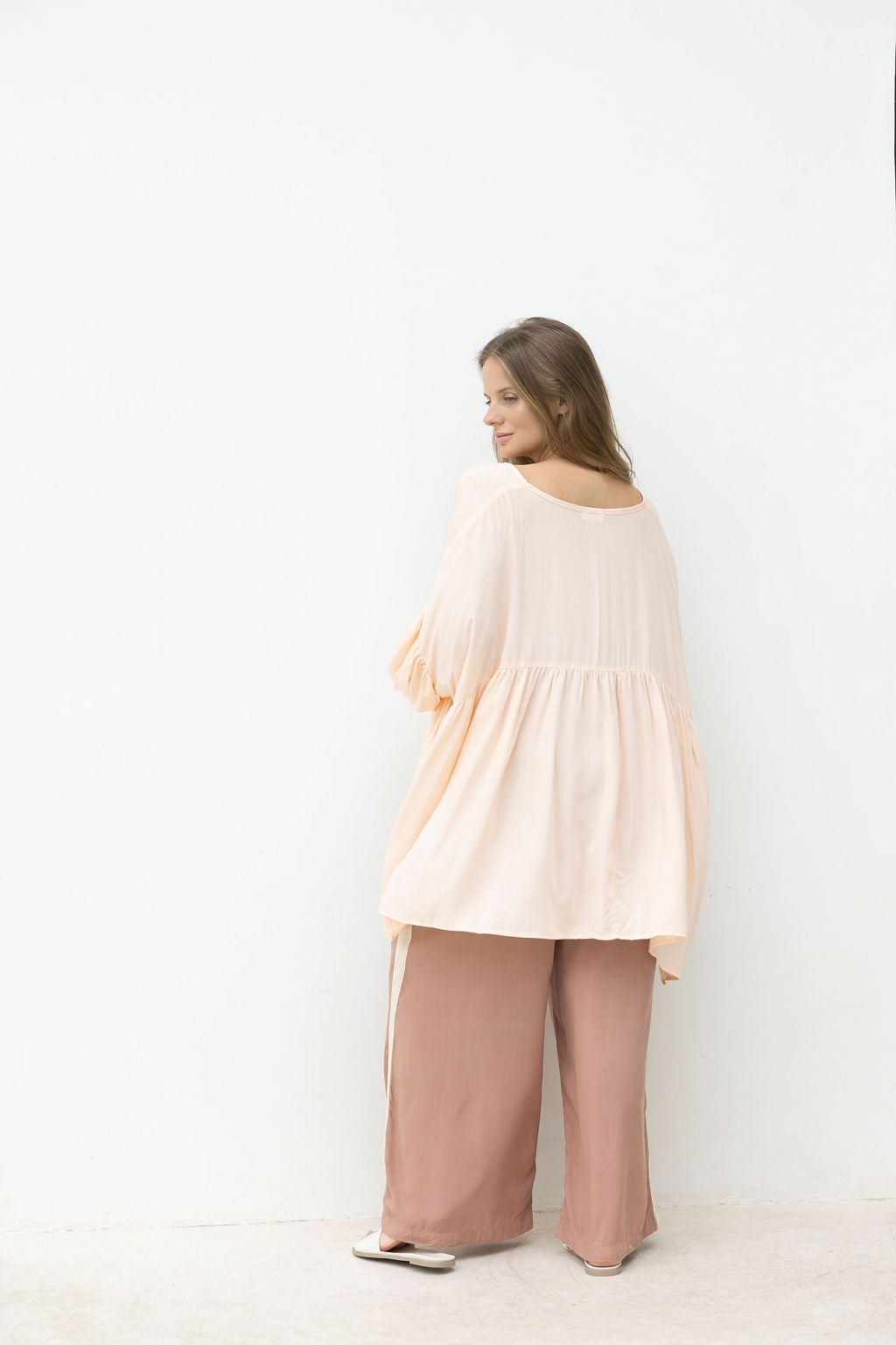 Woman wearing a blush top and wide-leg mocca coloured pants against a white coloured.