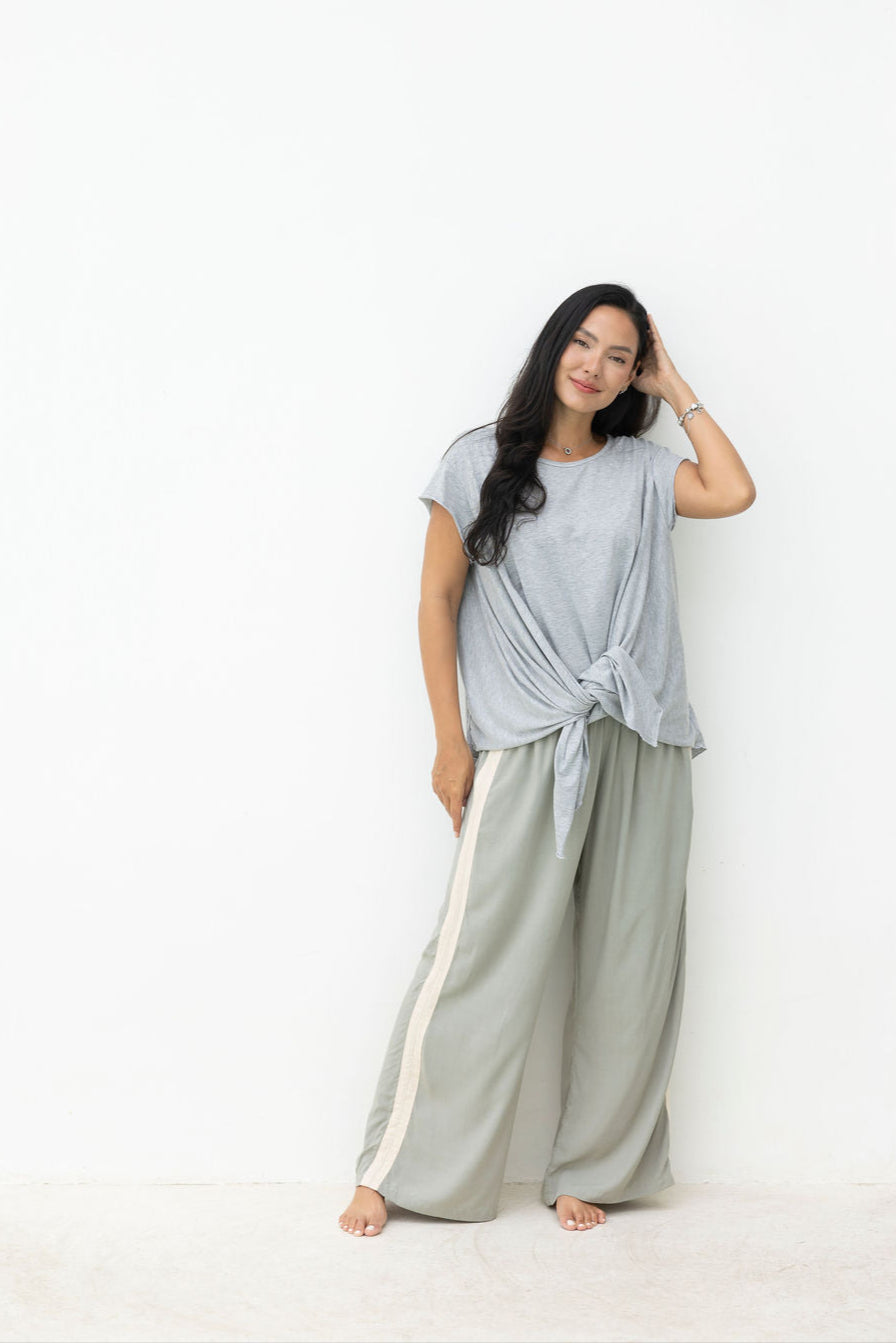 Woman wearing a light gray top and wide-leg pants with white stripes, standing against a white background.