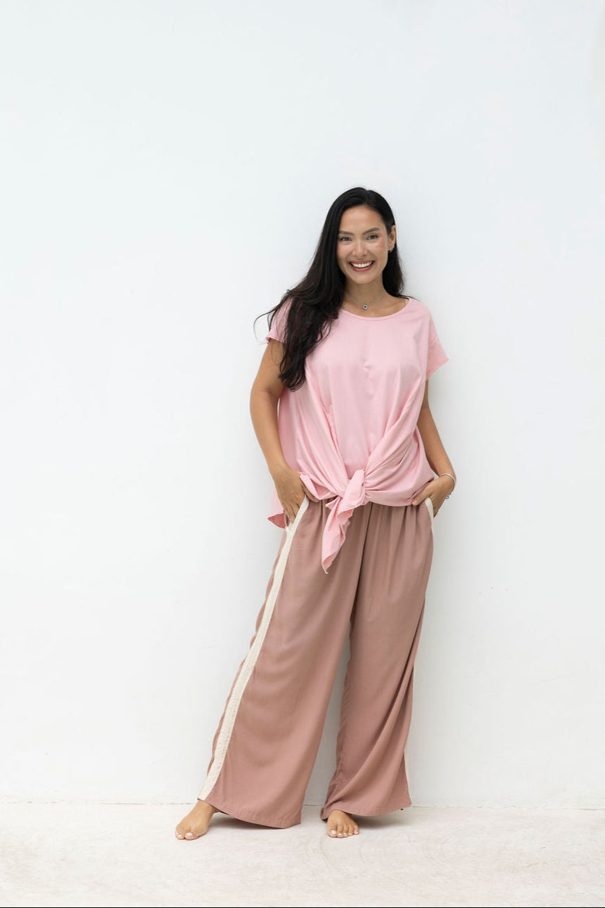 Woman wearing a pink top and beige pants with white stripes standing against a white background.