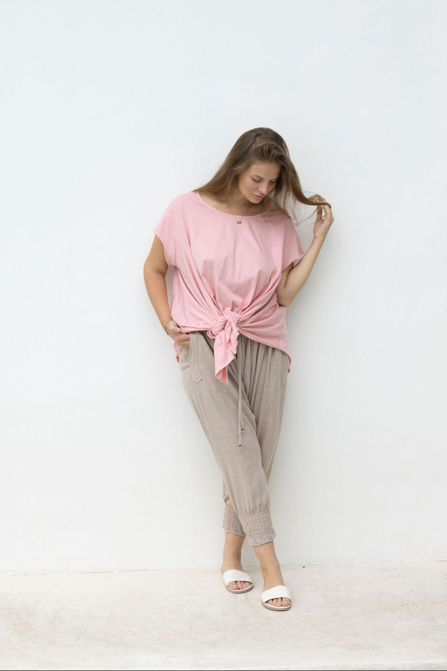 Woman wearing a pink top and beige pants against a white wall.