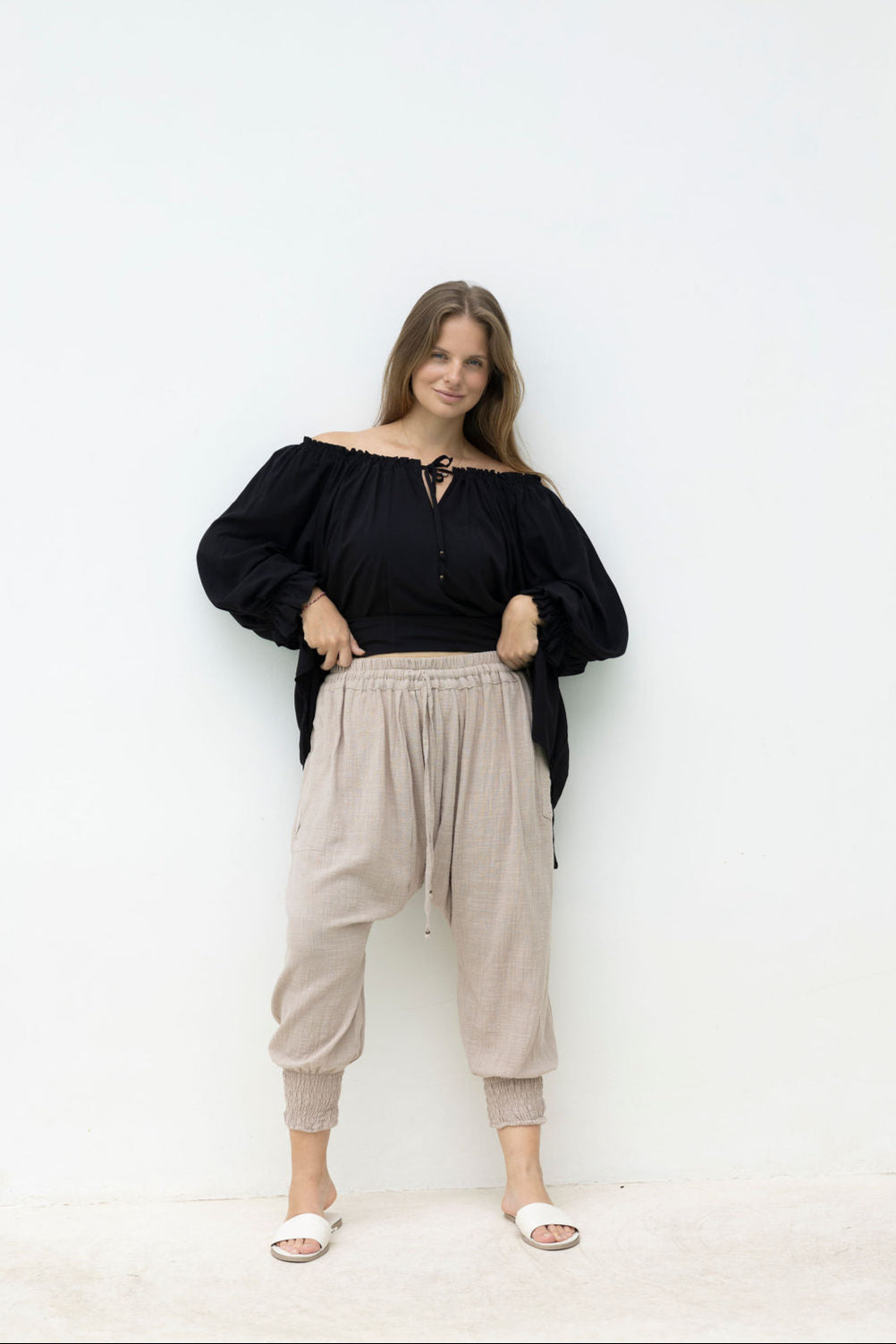 Woman wearing a black off-shoulder top and beige pants against a white background