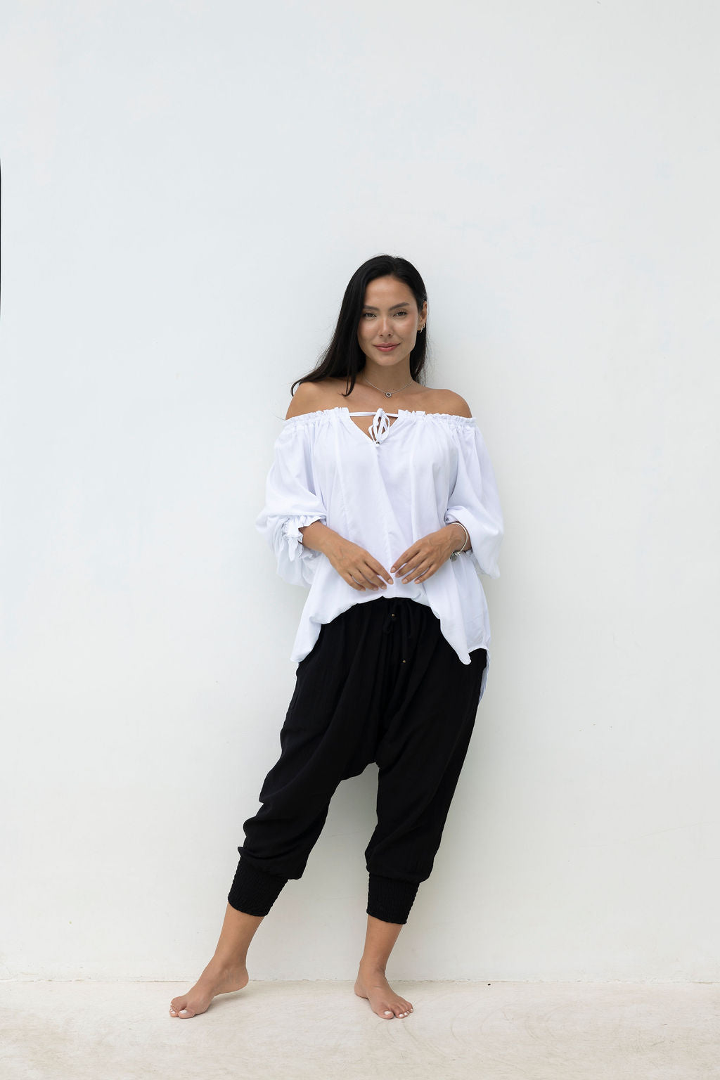 Woman wearing a white off-shoulder top and black harem pants against a white background