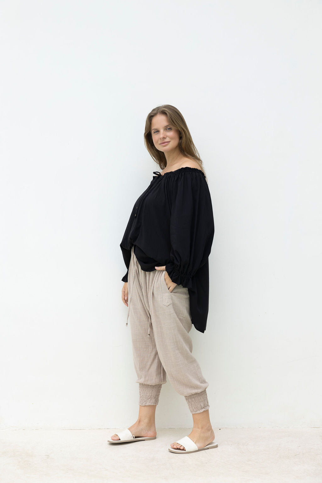 Woman wearing a black off-shoulder top and beige pants against a white background