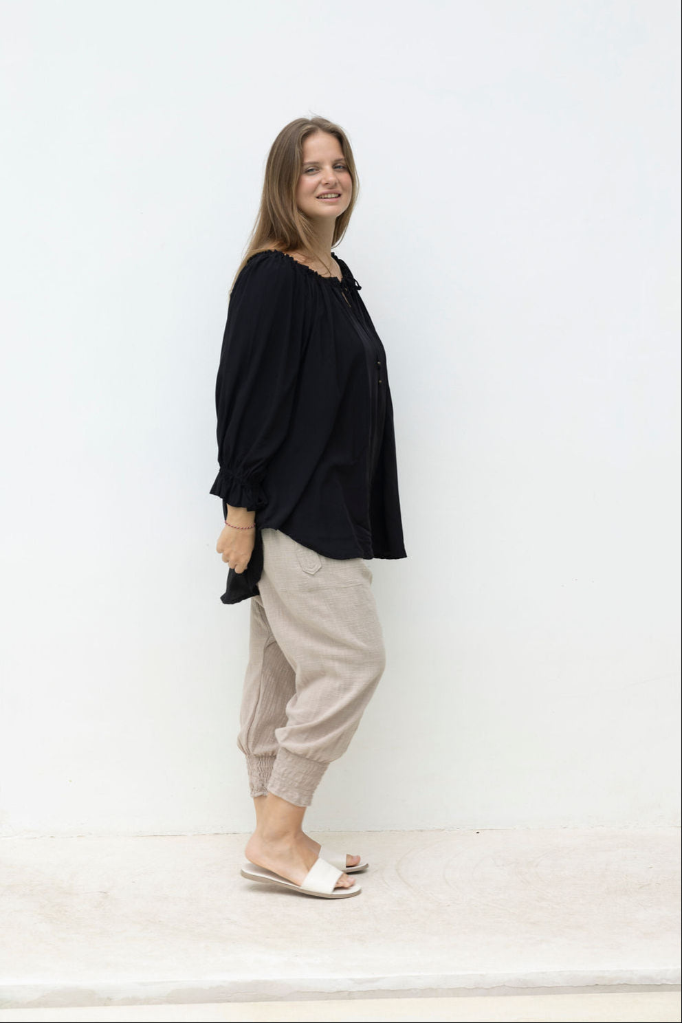 Woman wearing a black top and beige pants standing against a white wall.