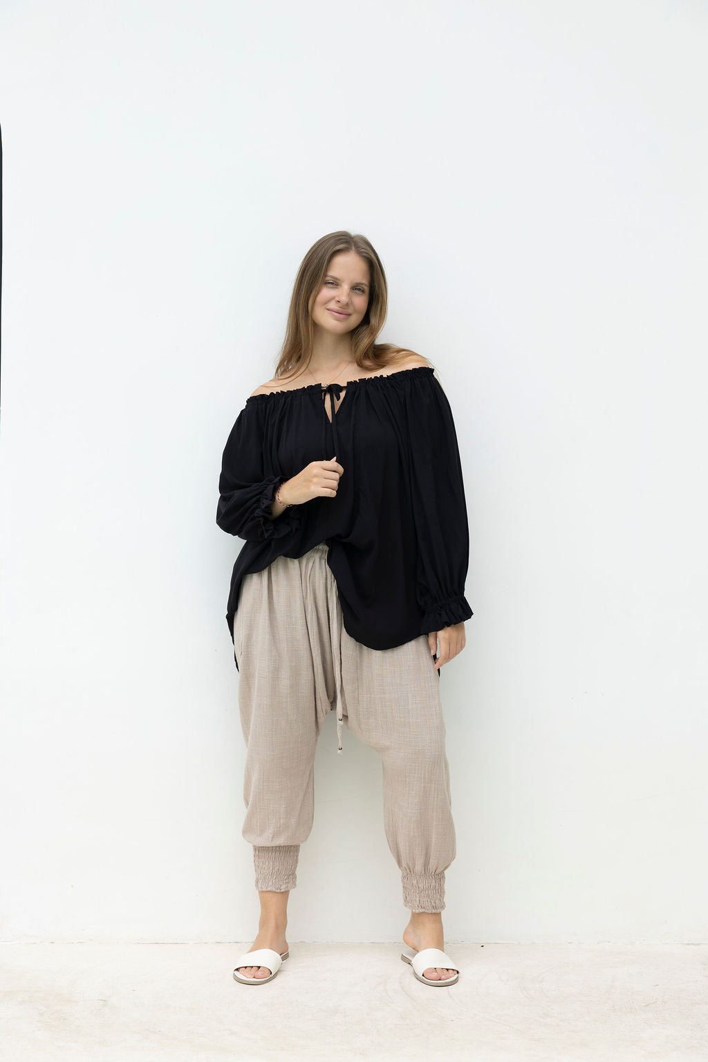 Woman wearing a black off-shoulder top and beige pants against a white background