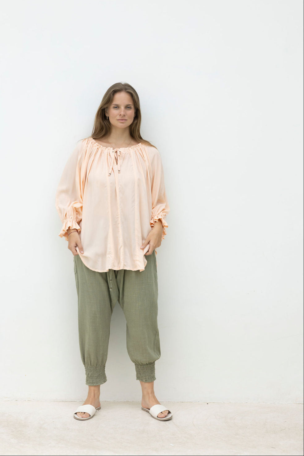 Woman wearing a light pink blouse and green pants against a white wall