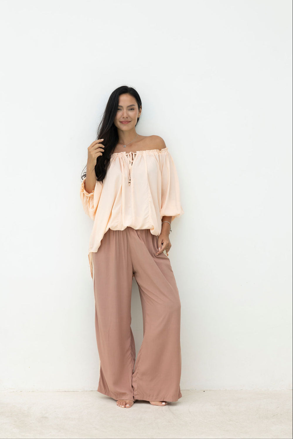 Woman wearing a light pink off-shoulder top and brown wide-leg pants on a white background