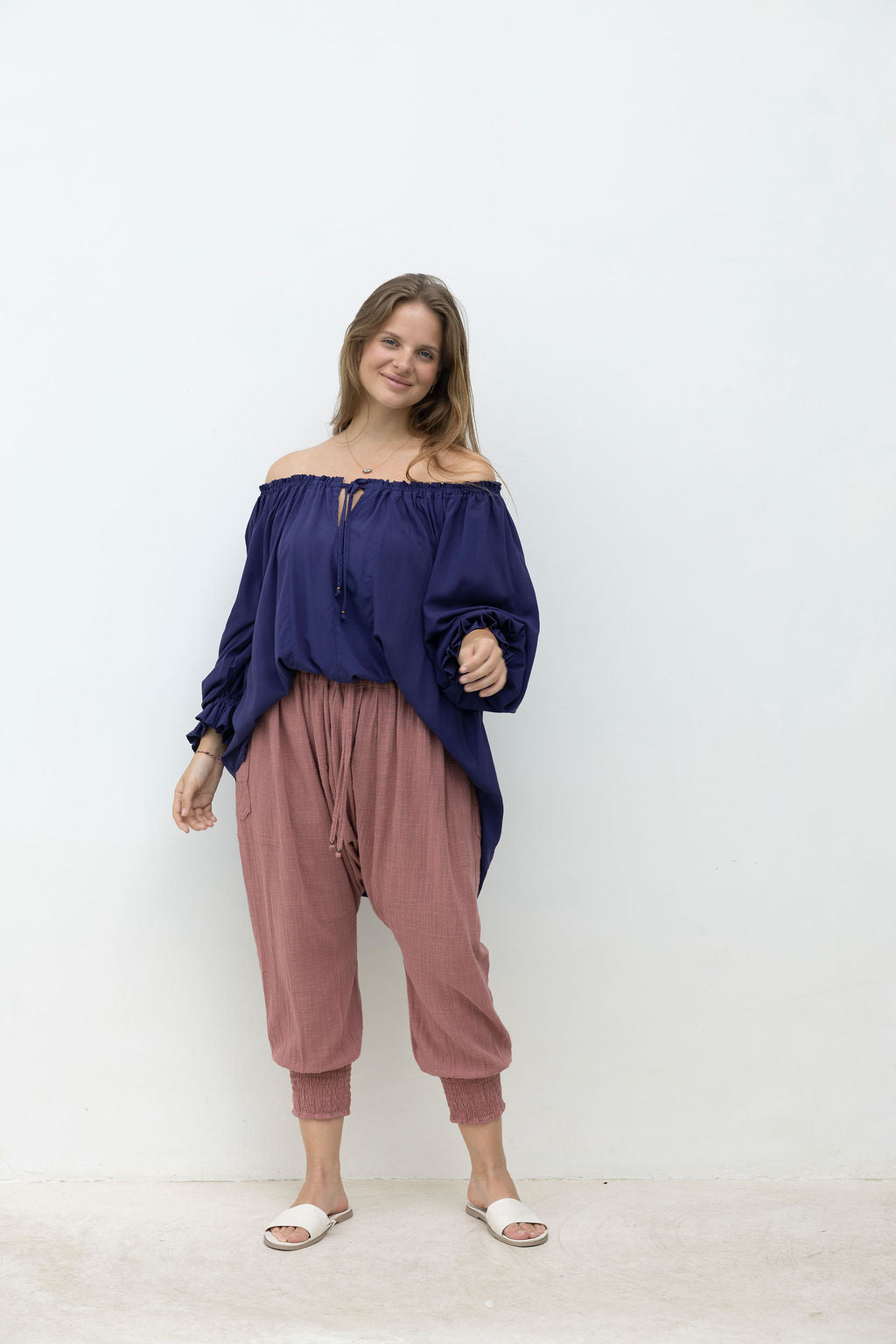 Woman wearing a blue off-shoulder top and pink pants against a white background