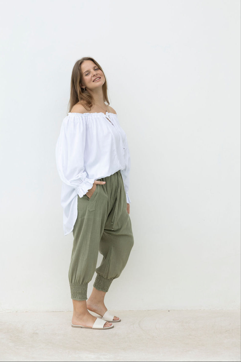 Woman wearing a white off-shoulder top and green pants against a white background