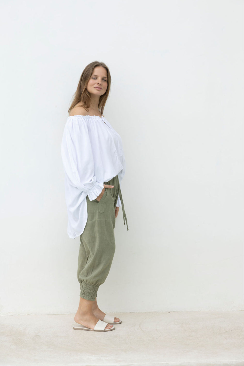 Woman wearing a white off-shoulder top and green pants against a plain background