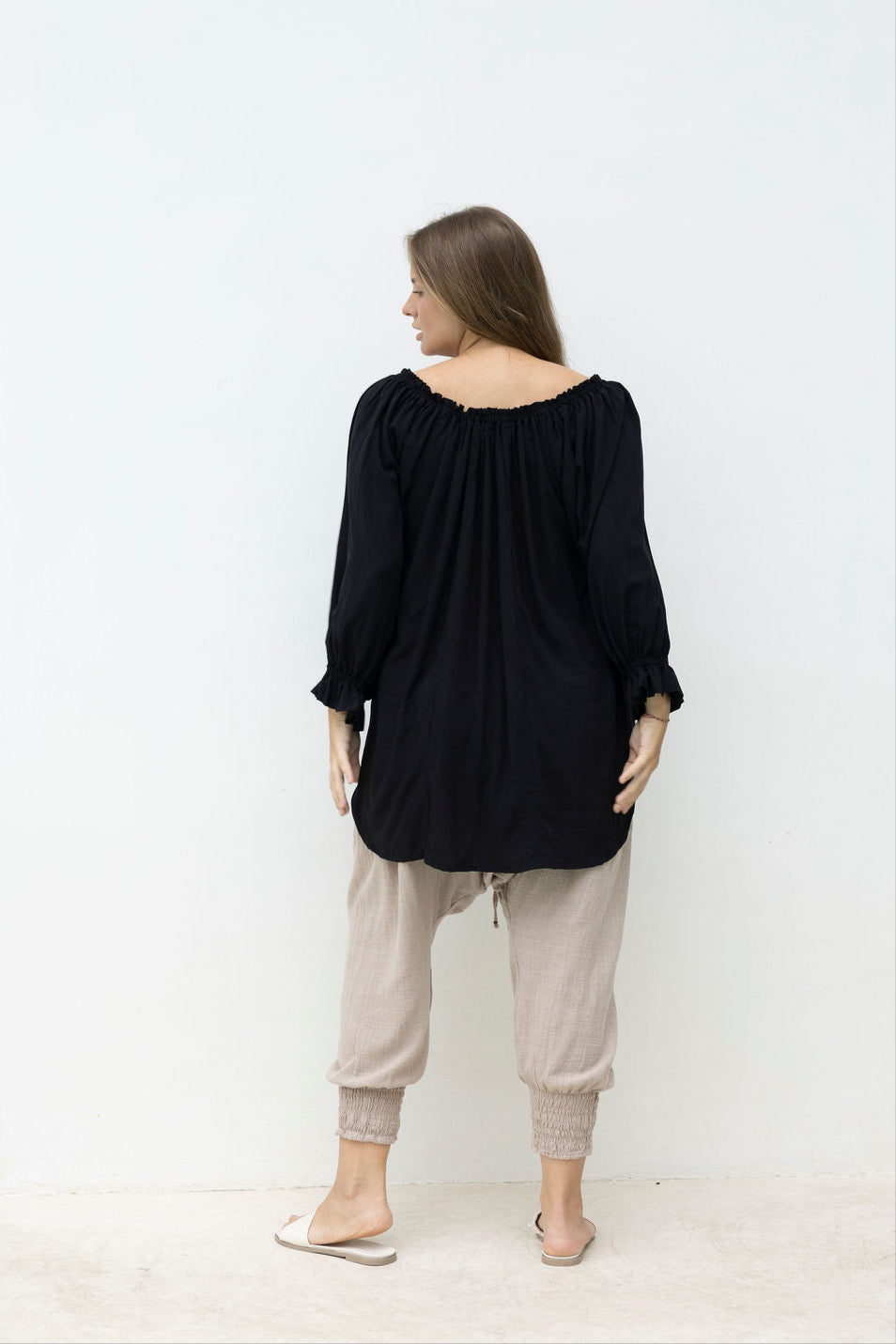 Woman wearing a black top and beige pants standing in front of a white wall with a tall mirror.
