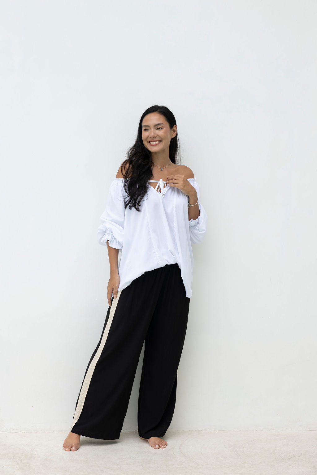 Woman wearing a white off-shoulder top and black pants with a stripe against a white background