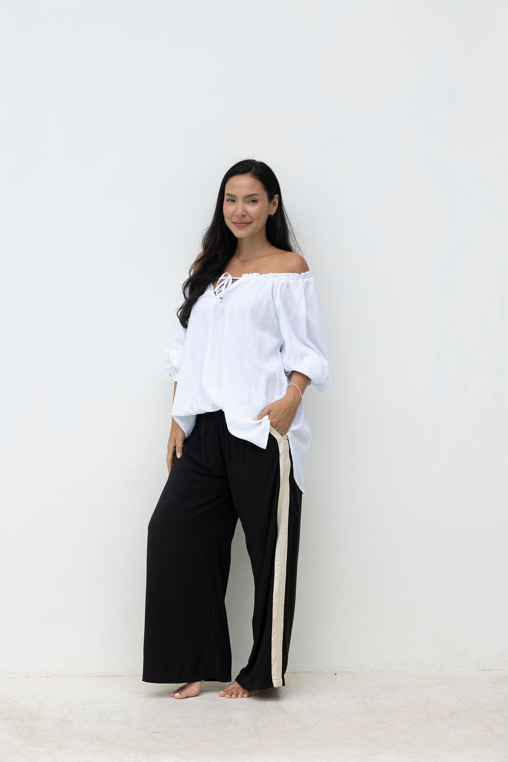 Woman wearing a white off-shoulder top and black pants against a white background
