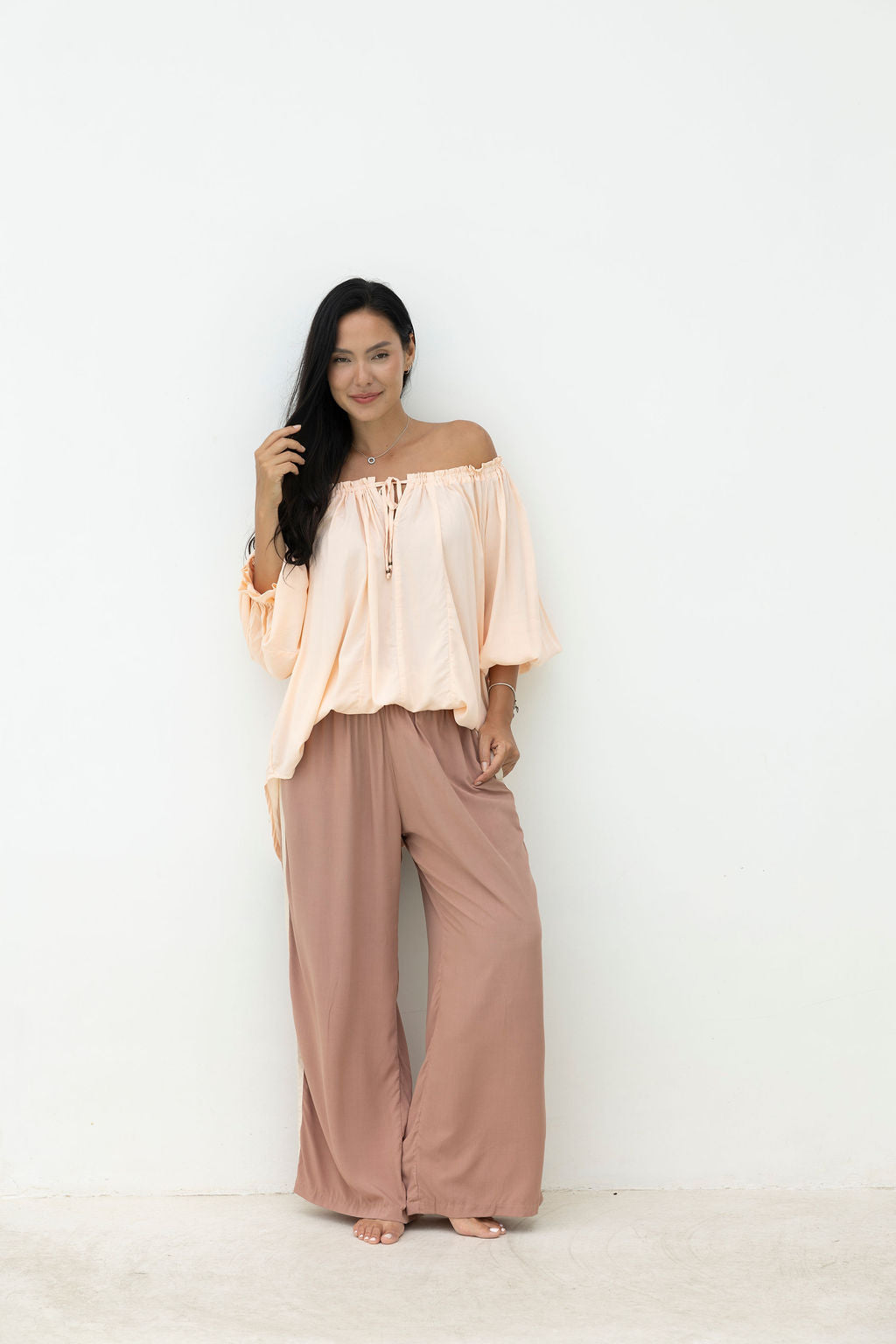 Woman wearing a peach off-shoulder top and brown wide-leg pants against a white background