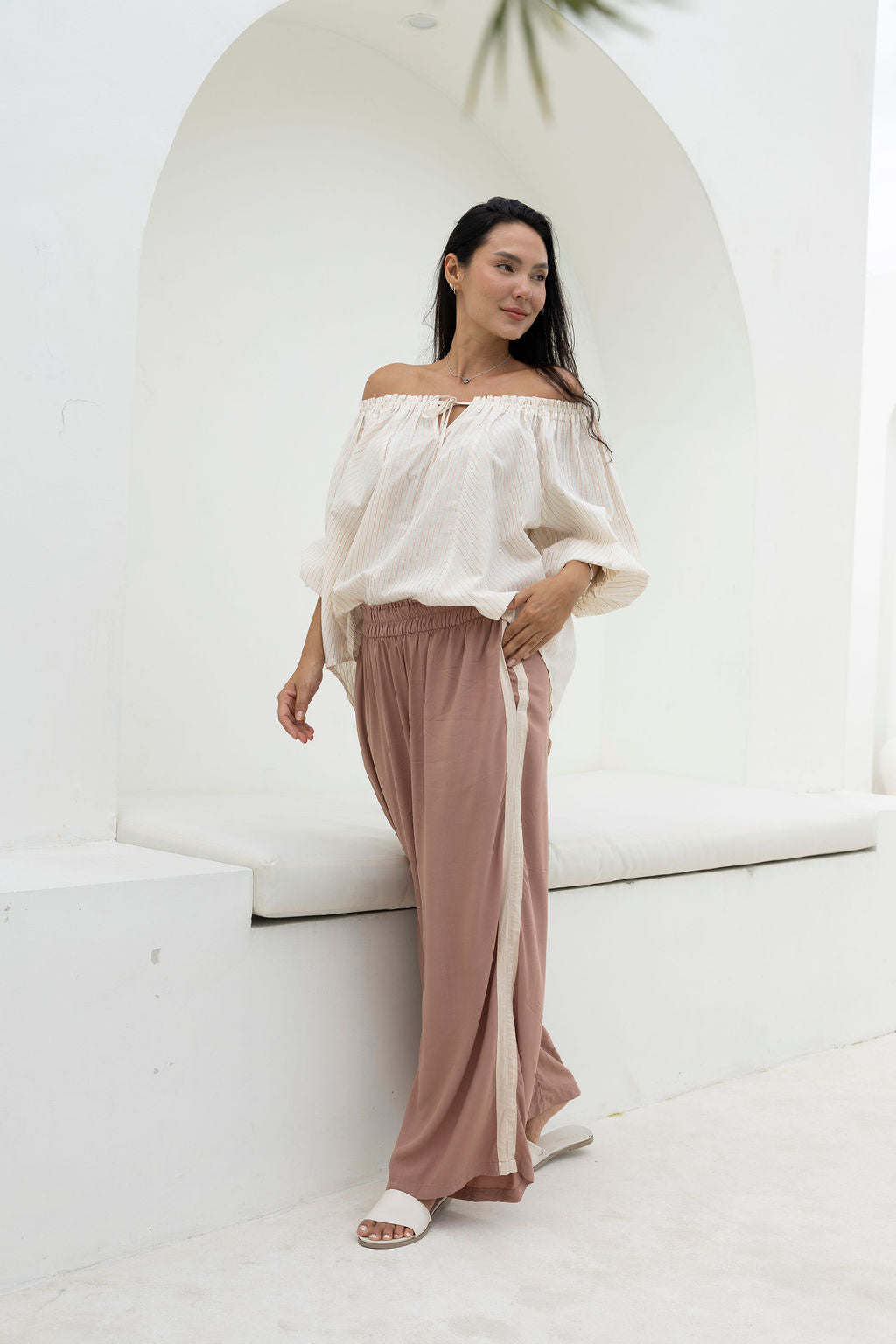 Woman wearing a white off-shoulder top and pink pants in a minimalistic setting.