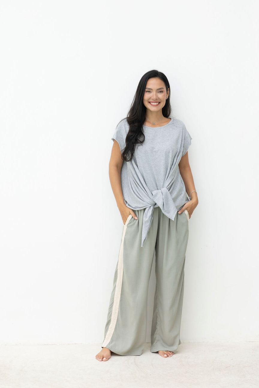 Woman wearing a gray top and light green pants standing against a white wall.