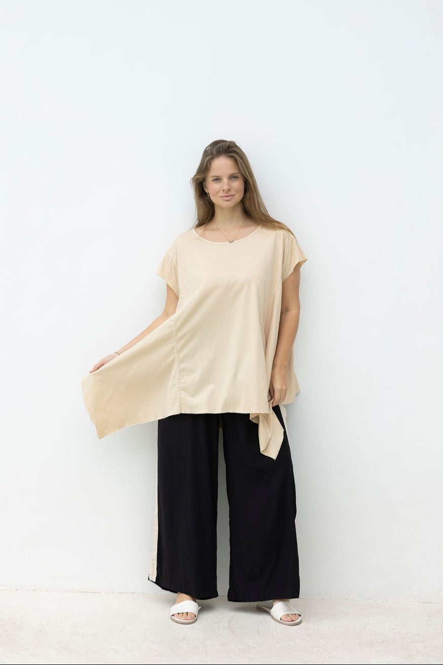 Woman wearing a beige top and black pants standing against a white wall.