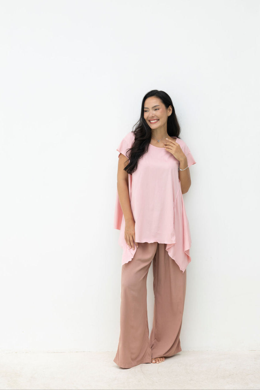 Woman wearing a pink top and brown pants standing against a white wall.