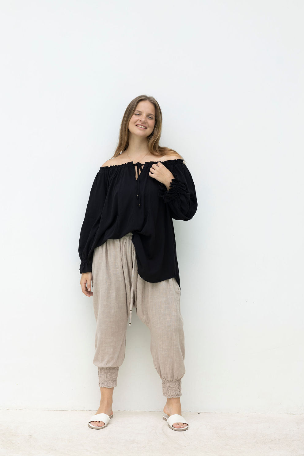 Woman wearing a black off-shoulder top and beige pants against a white background