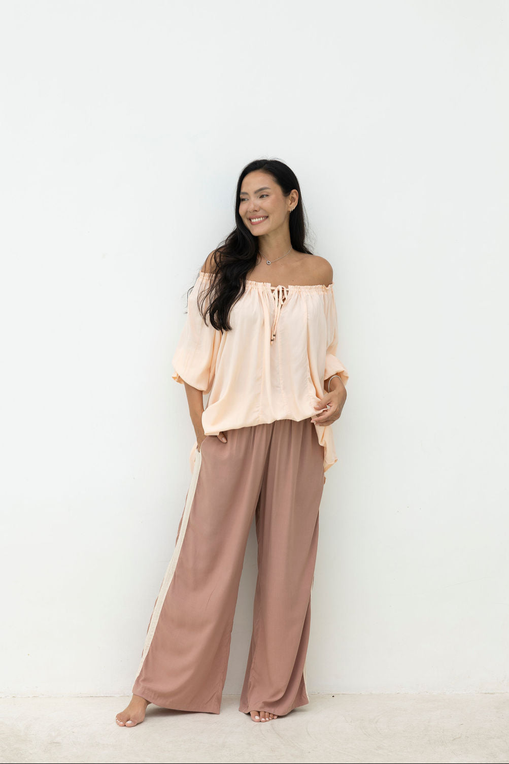 Woman wearing a light pink off-shoulder top and brown wide-leg pants on a white background