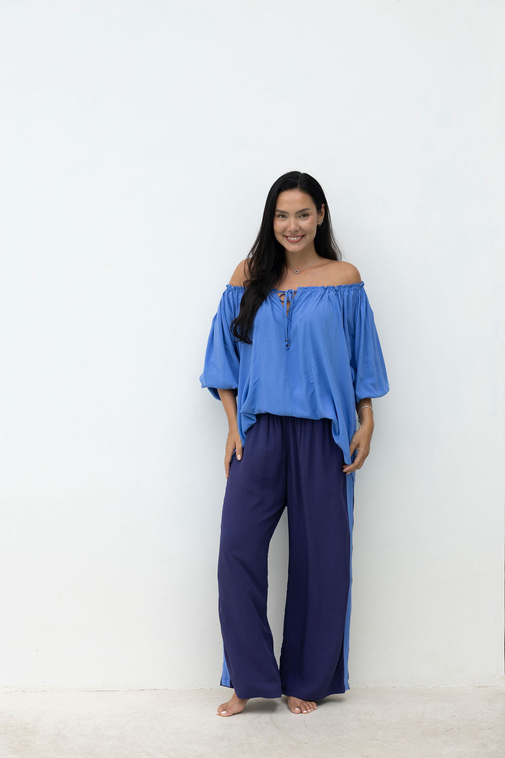 Woman wearing a blue off-shoulder top and purple pants against a white background