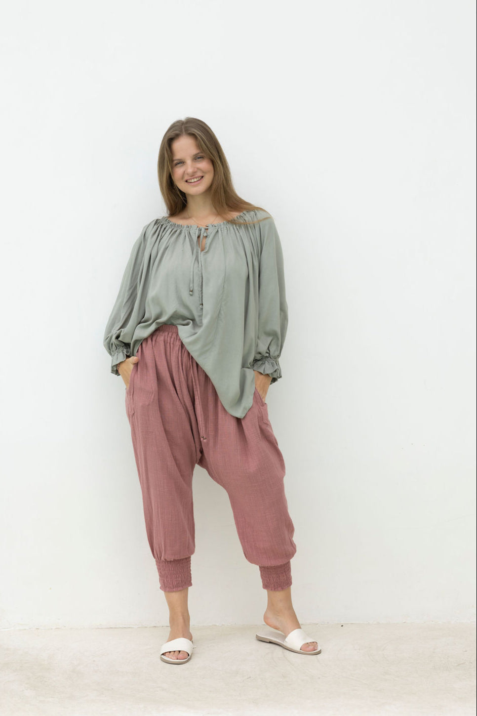 Woman wearing a green top and pink pants against a white wall