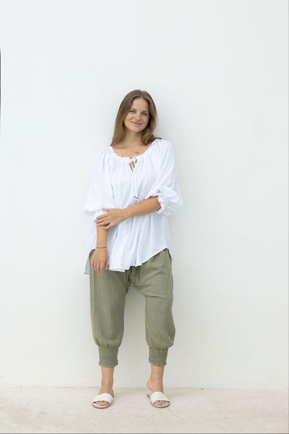 Woman wearing a white blouse and green pants standing against a white wall.