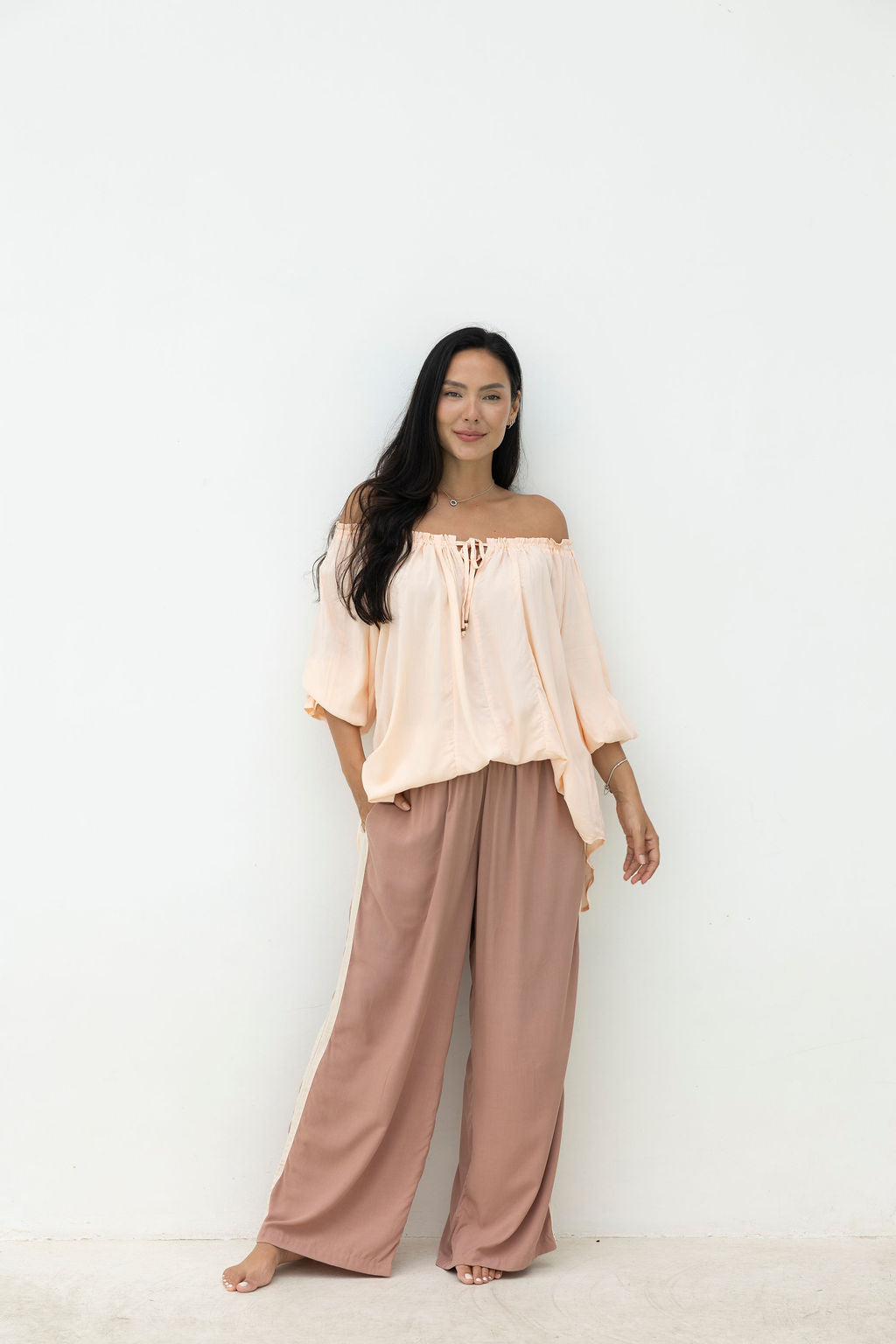 Woman wearing a light pink off-shoulder top and brown wide-leg pants against a white background