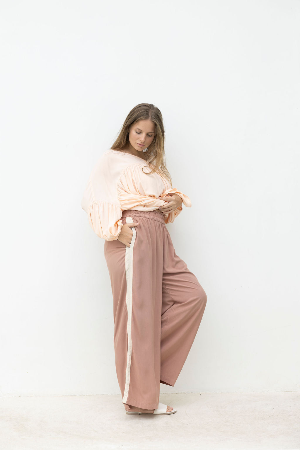Woman wearing a light pink top and brown wide-leg pants against a white background